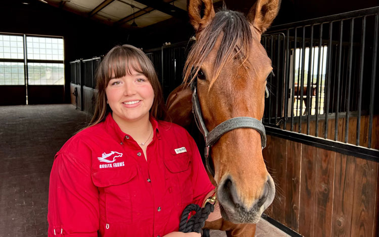 Abby wearing a red rosita farms shirt and posing with a young horse in the stalls.